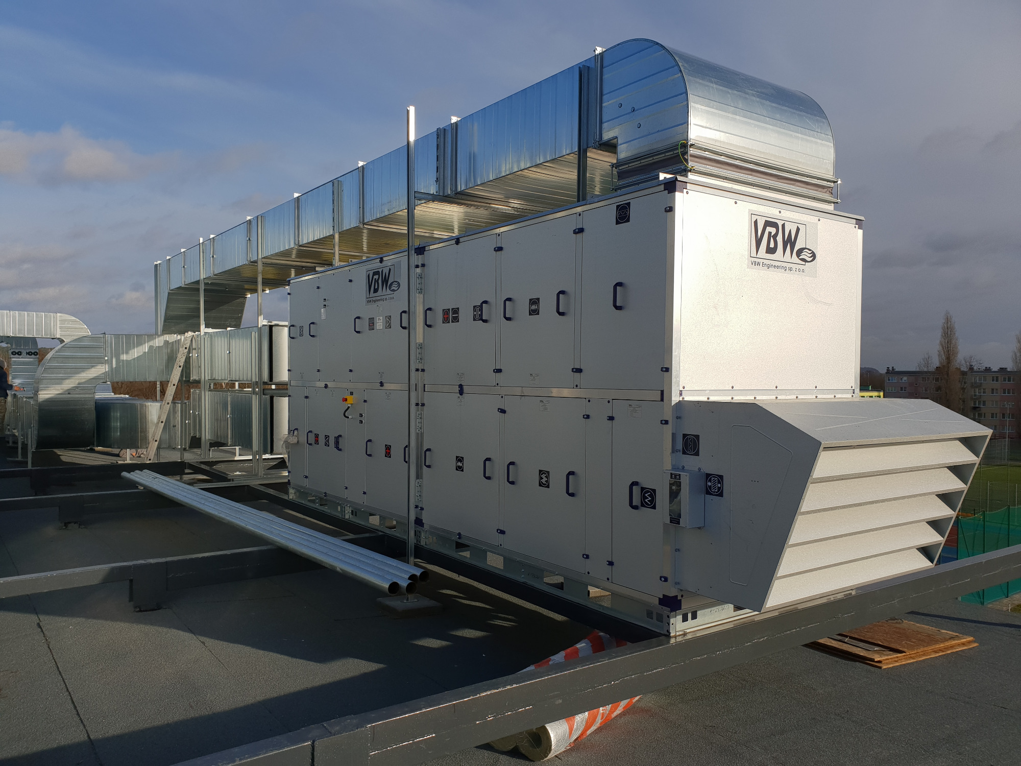 Zewnętrzna instalacja centrali wentylacyjnej VBW z kanałami na dachu budynku, widoczne elementy mocujące i zabezpieczenia.
