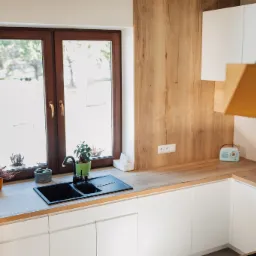 Jasna kuchnia z drewnianym blatem, czarnym zlewem pod oknem i białymi szafkami, uzupełniona o rośliny w doniczkach i retro radio.