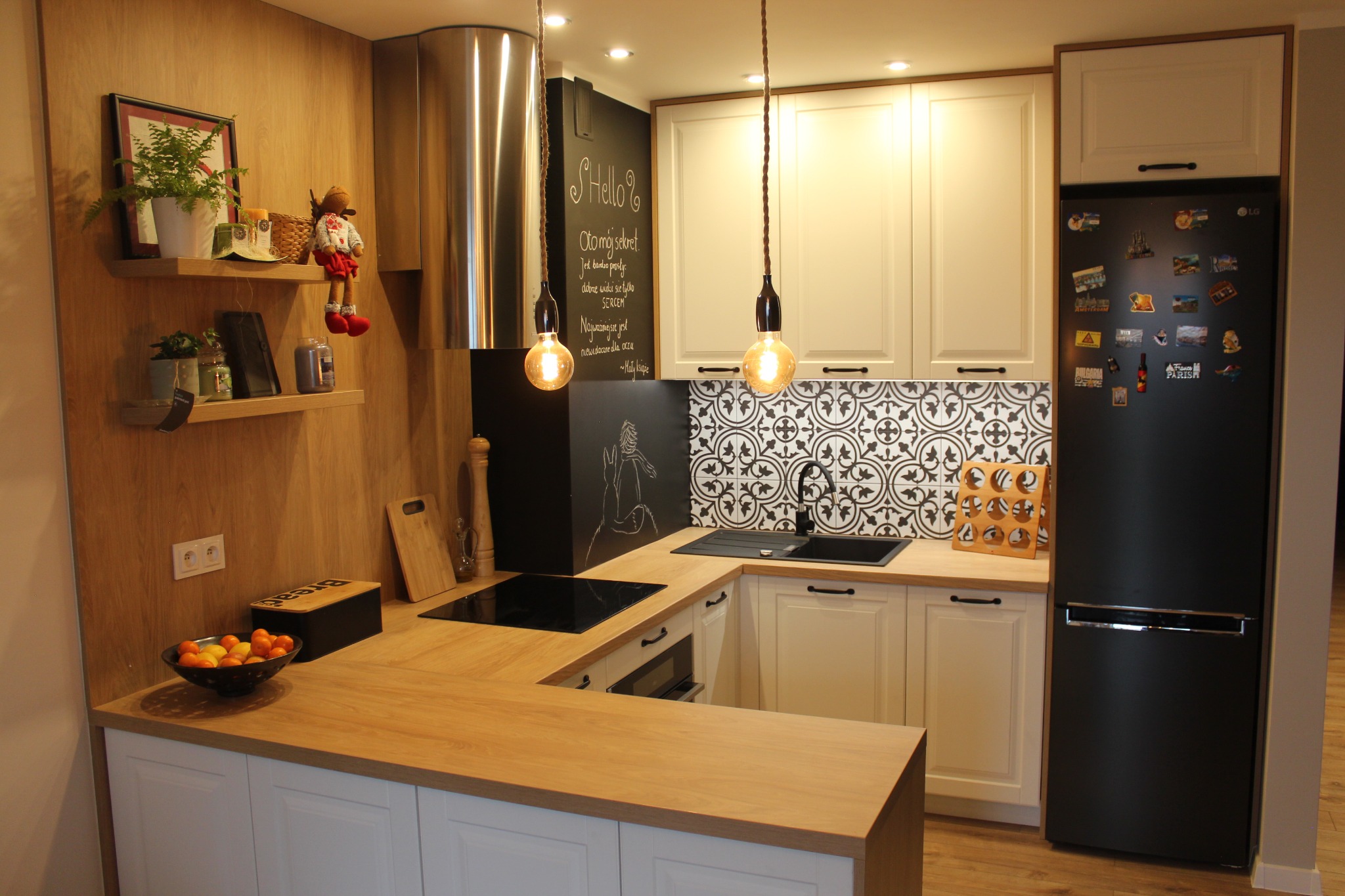 Jasna kuchnia w stylu klasycznym z drewnianym blatem, czarnym zlewem i płytkami w marokański wzór, oświetlona wiszącymi lampami z żarówkami Edisona, z czarną lodówką ozdobioną magnesami i tablicą...