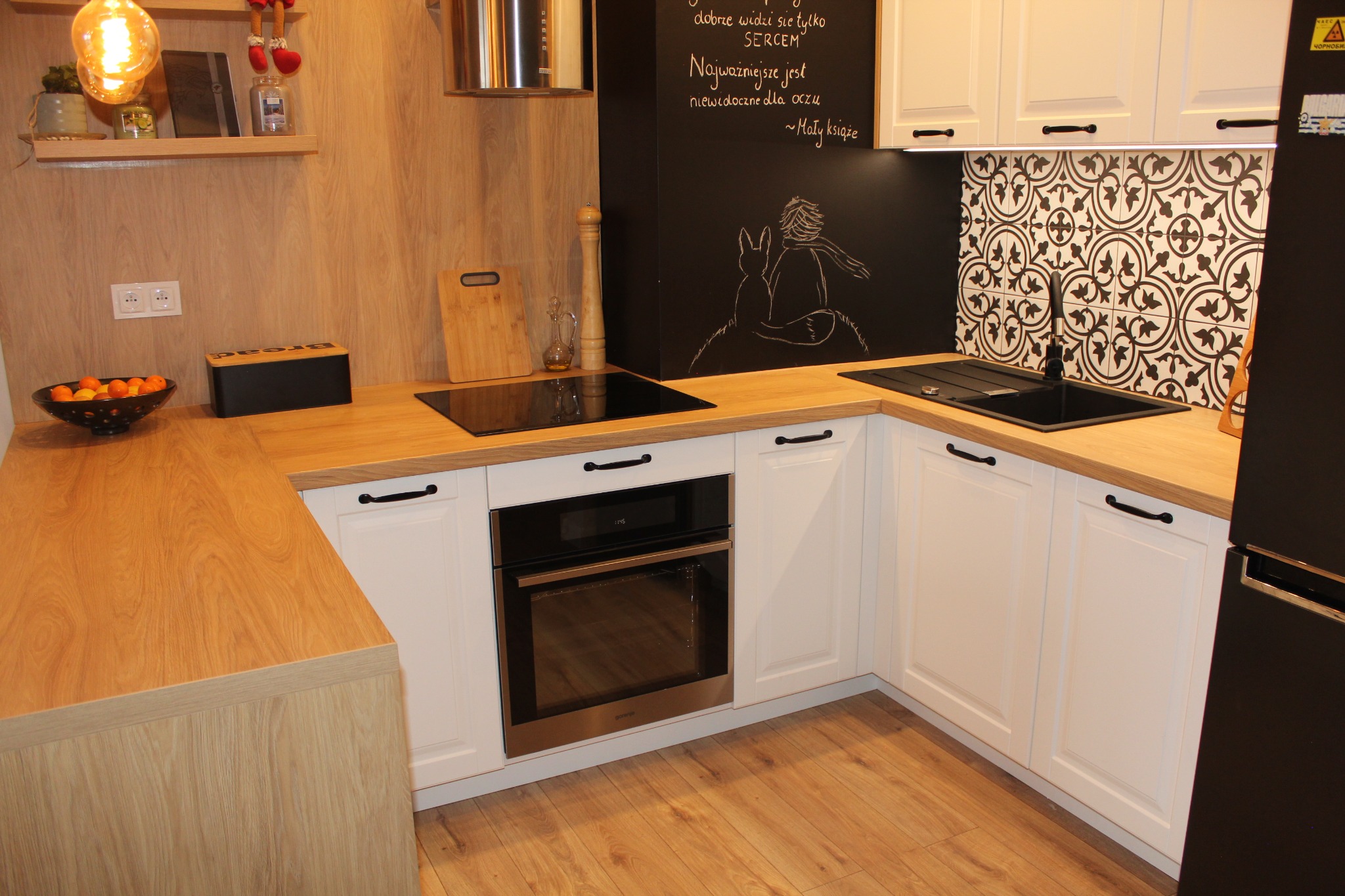 Nowoczesna kuchnia z białymi szafkami, drewnianym blatem, czarnym zlewem i czarną ścianą z rysunkiem Małego Księcia.