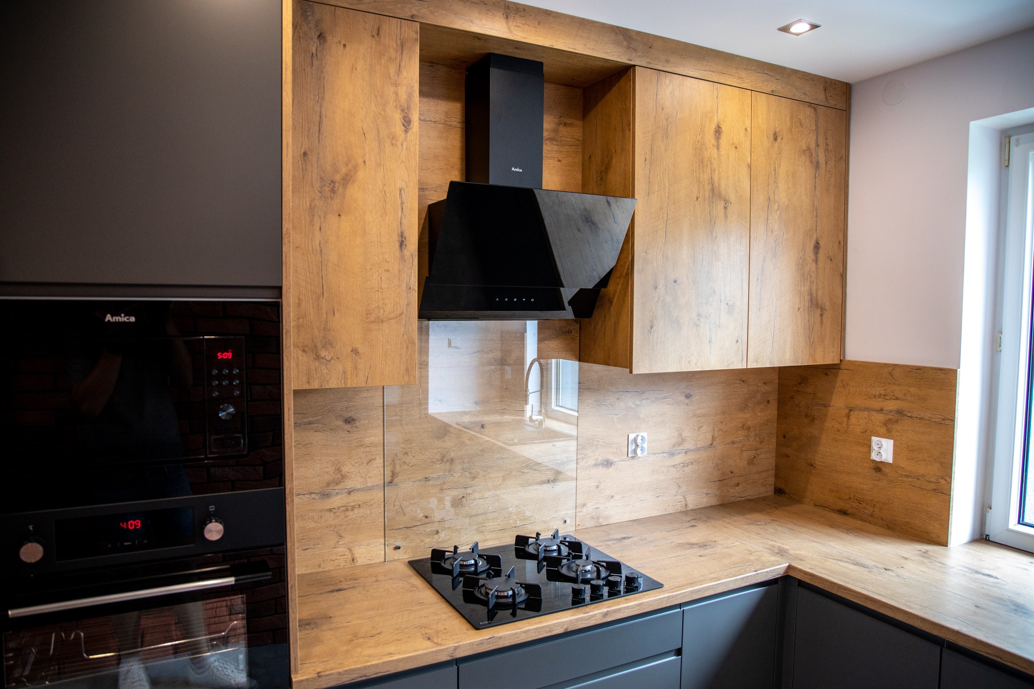 Nowoczesna kuchnia z drewnianymi frontami szafek wiszących i szarymi szafkami dolnymi, czarnym okapem i płytą gazową, z piekarnikiem Amica w zabudowie.