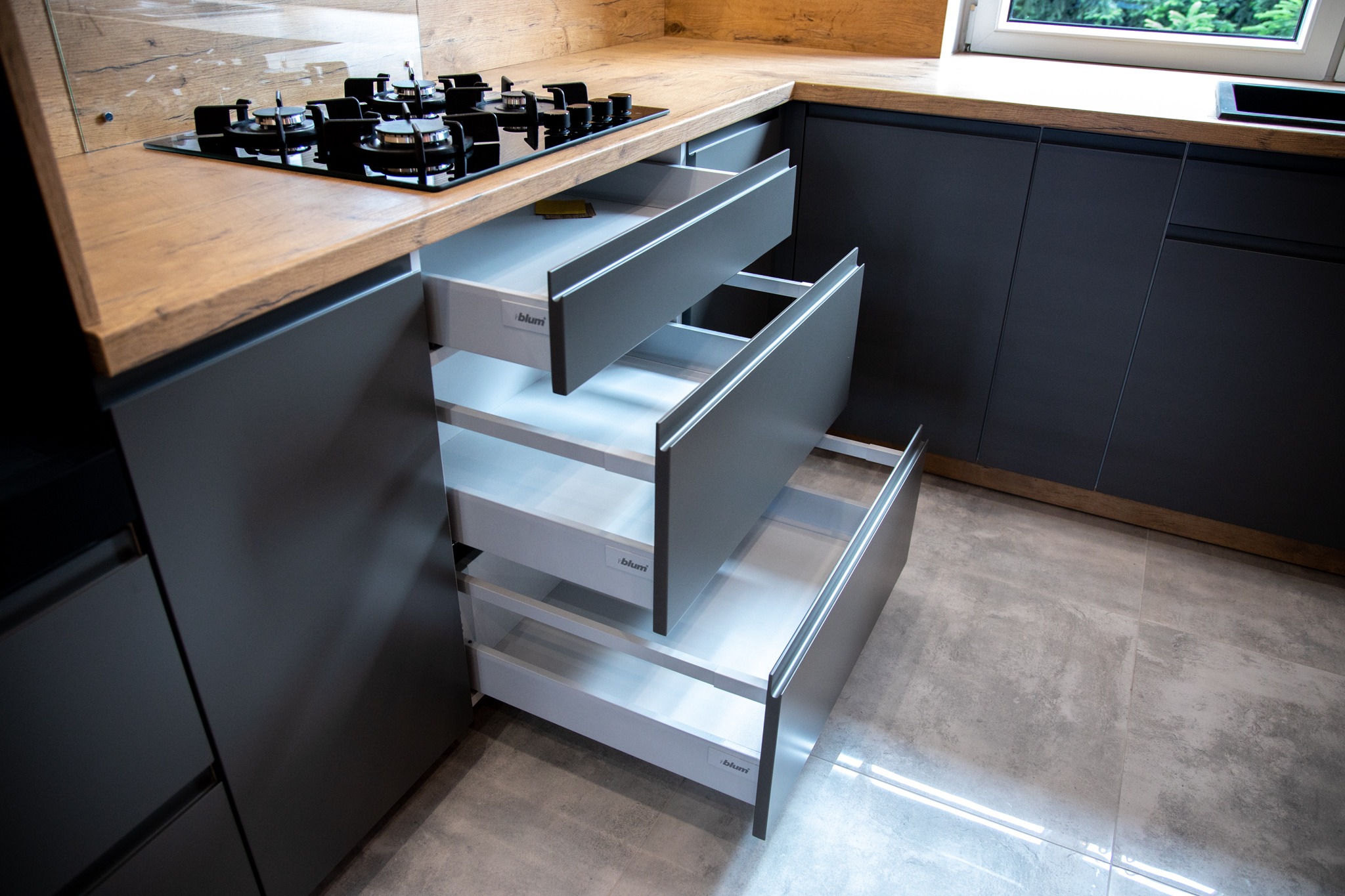 Szare szafki kuchenne z drewnianym blatem i otwartymi szufladami z systemem Blum, widoczne od frontu, z płytą gazową na blacie.