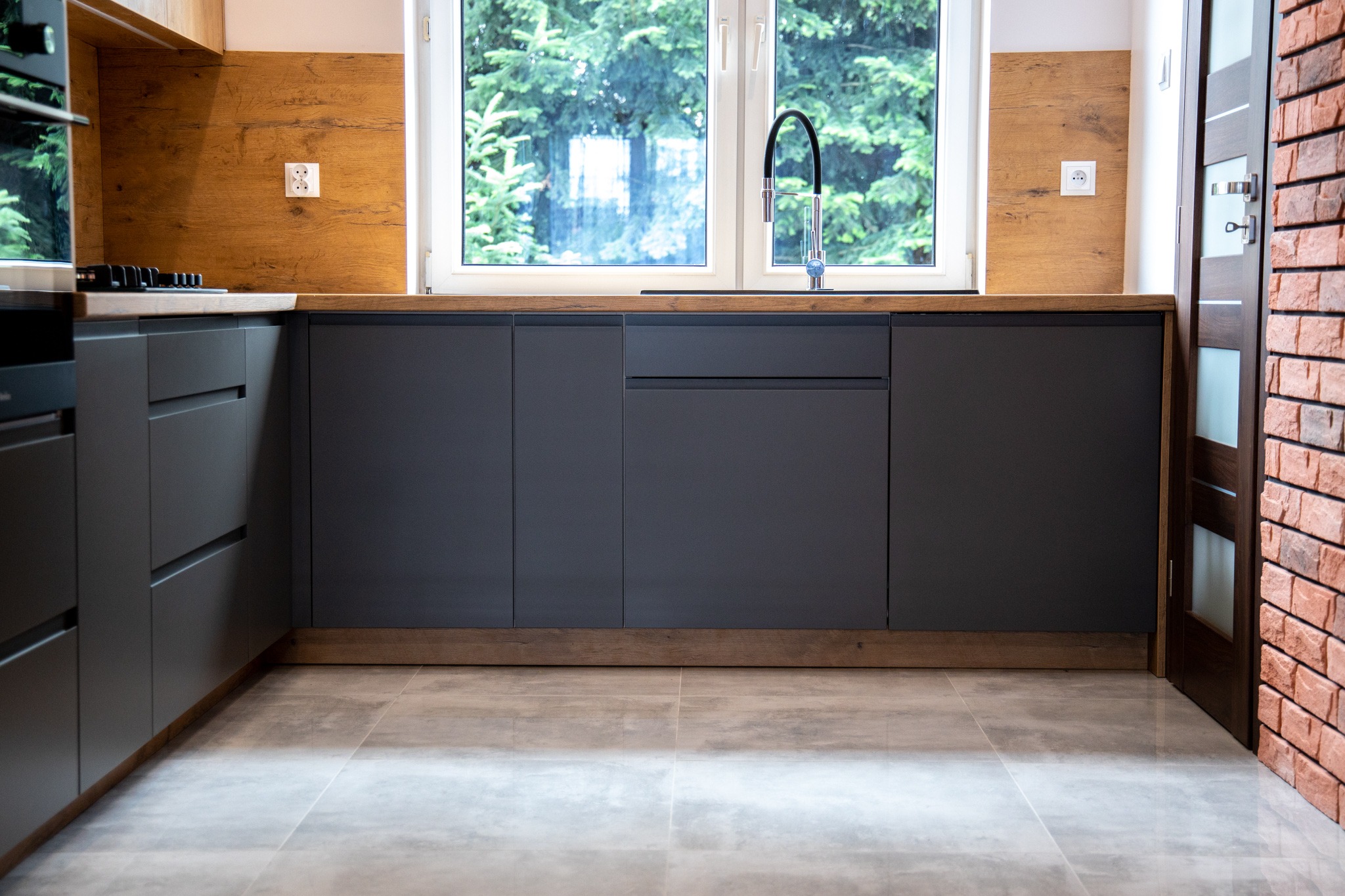Nowoczesna kuchnia z szarymi frontami szafek, drewnianym blatem i oknem z widokiem na zieleń, wykończona ceglaną ścianą i płytkami podłogowymi w odcieniach szarości.