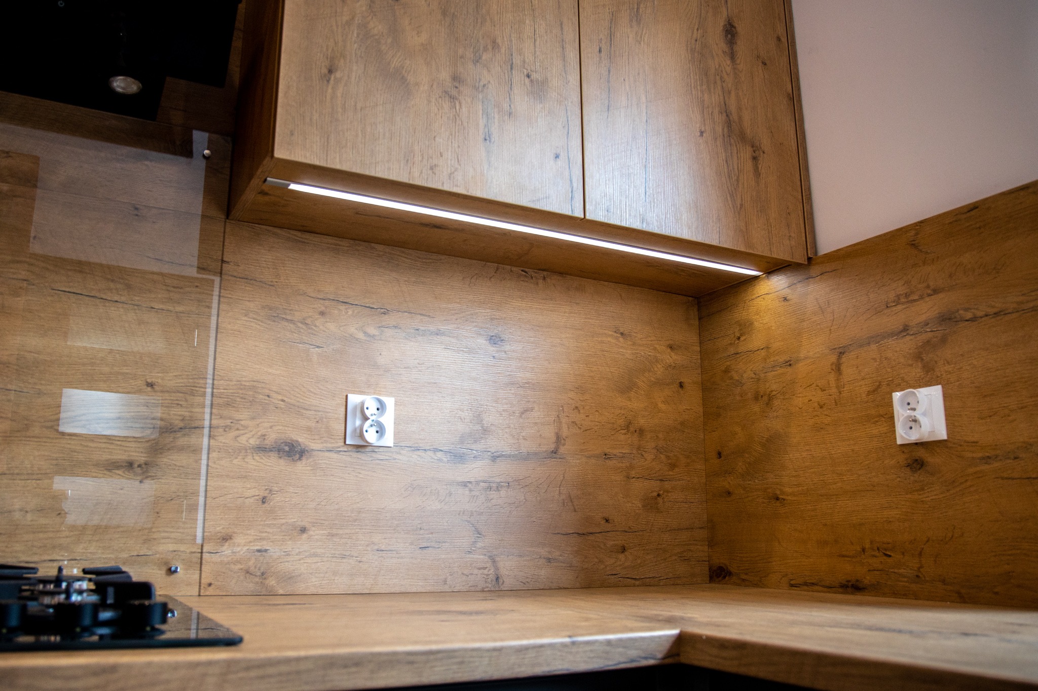 Fragment wykończonej kuchni z drewnopodobnymi frontami szafek, blatem i panelem ściennym, podświetlona listwą LED pod szafką wiszącą, z widocznymi gniazdkami elektrycznymi i fragmentem płyty...