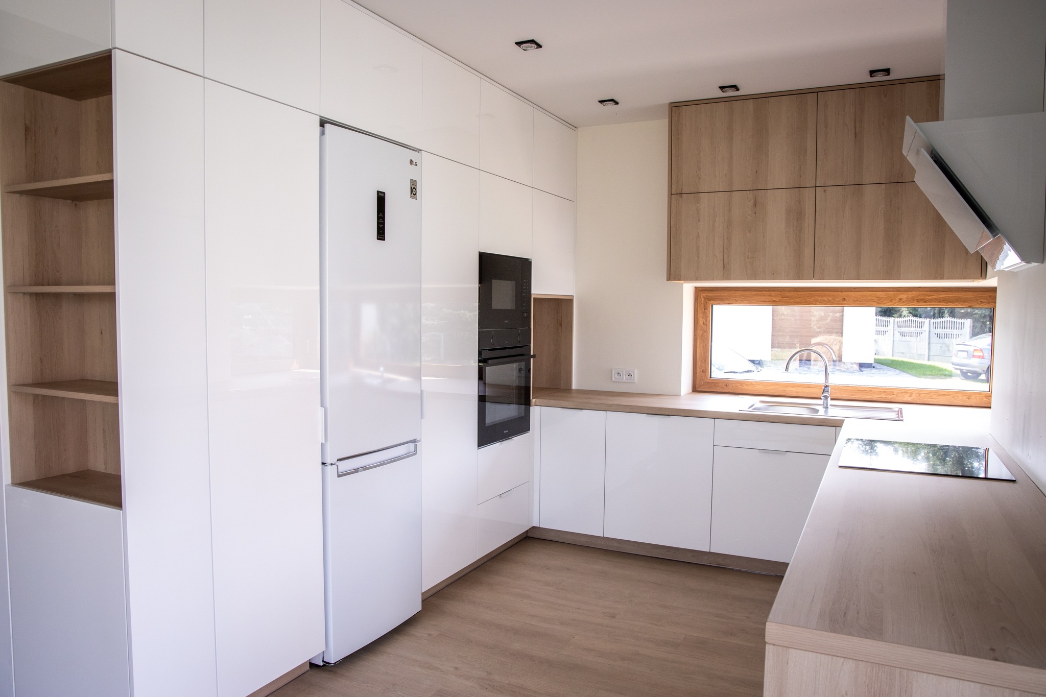 Nowoczesna kuchnia z białymi szafkami na wysoki połysk i drewnianymi akcentami, widok na okno z widokiem na ogród.