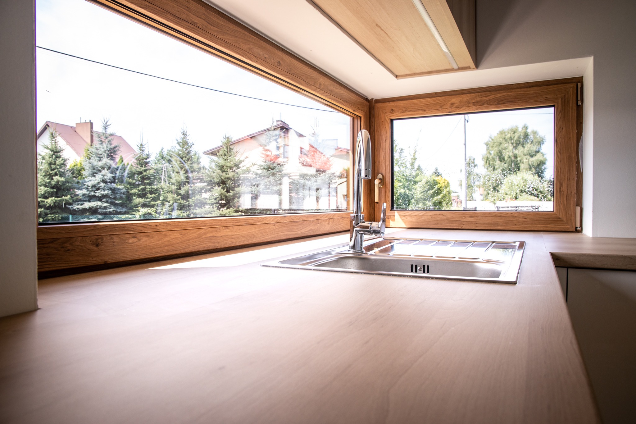Jasny drewniany blat z wbudowanym zlewozmywakiem i baterią, duże okno z widokiem na dom i drzewa w tle.