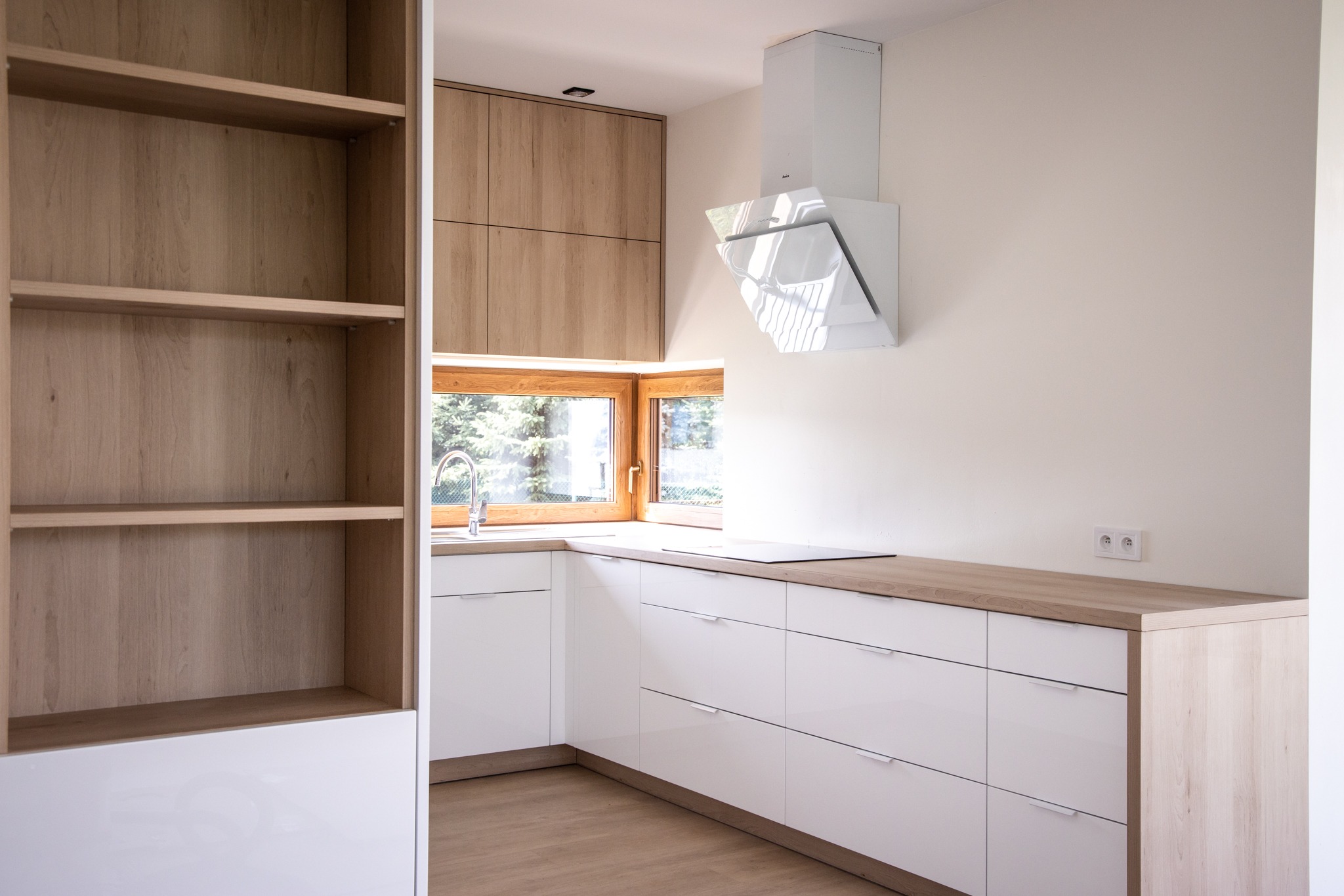 Jasna kuchnia w stylu skandynawskim z białymi frontami szafek i drewnianym blatem, z widokiem na zieleń za oknem i nowoczesnym okapem.