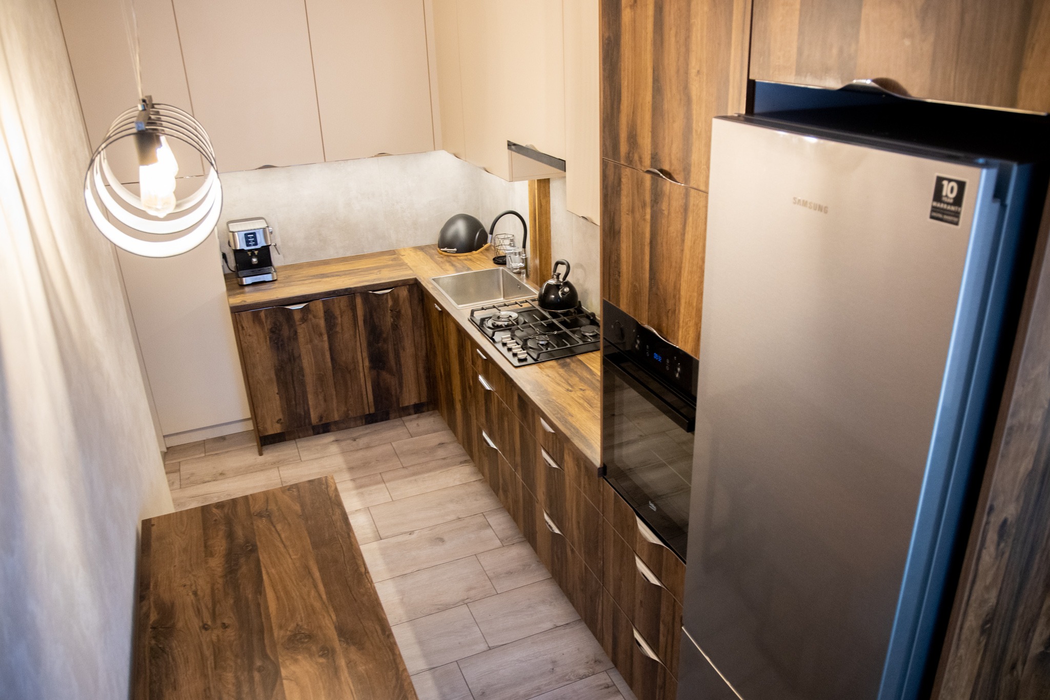 Nowoczesna kuchnia z drewnianymi frontami szafek i beżowymi górnymi szafkami, oświetlona designerską lampą wiszącą, z widocznym ekspresem do kawy i czajnikiem.