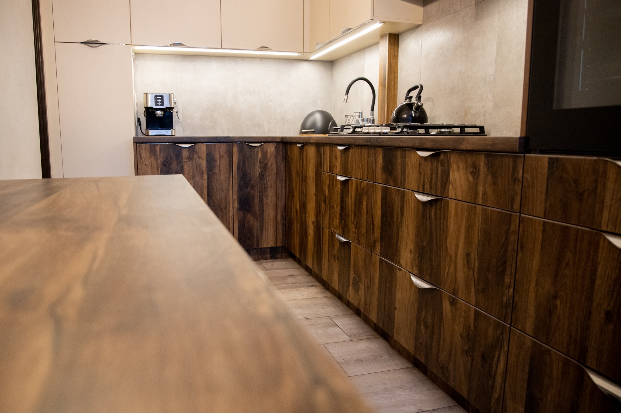 Fragment zabudowy kuchennej z drewnopodobnymi frontami szafek i szuflad, widoczny ekspres do kawy, czajnik na płycie grzewczej i blat stołu na pierwszym planie.