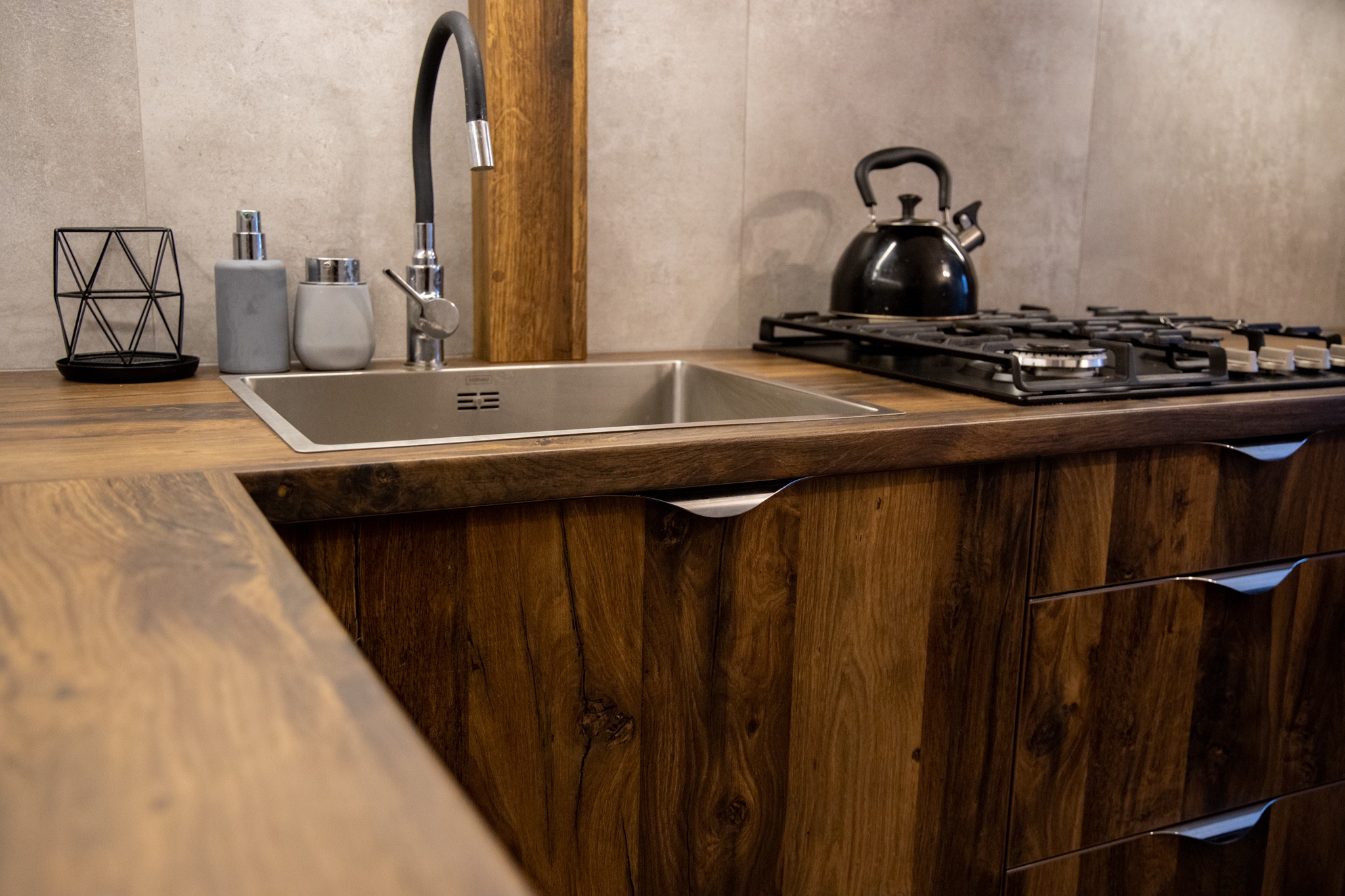 Fragment blatu kuchennego z drewnianym wykończeniem, stalowy zlew z czarną baterią, czajnik na płycie grzewczej, dekoracyjne pojemniki na blacie, szare płytki na ścianie.
