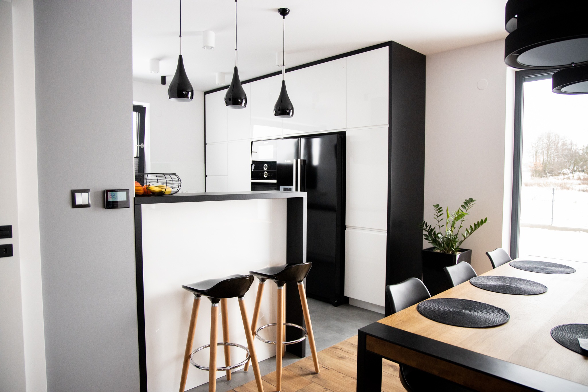 Nowoczesna kuchnia z białymi szafkami, czarnym sprzętem AGD i barkiem z hokerami na drewnianych nogach, widok na jadalnię z drewnianym stołem i czarnymi podkładkami.