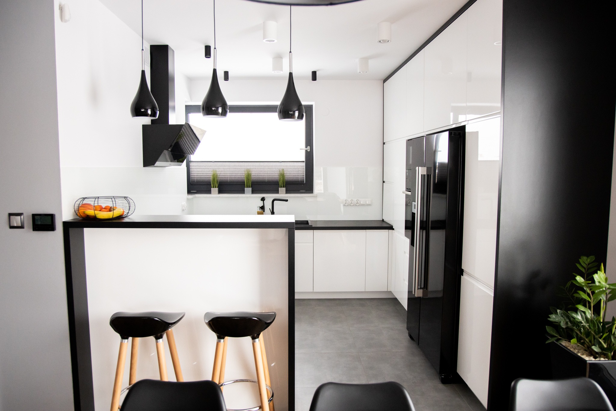 Nowoczesna kuchnia w bieli i czerni z wyspą, oświetlona wiszącymi lampami w kształcie kropli, z drewnianymi hokerami i owocami w drucianym koszu na blacie.