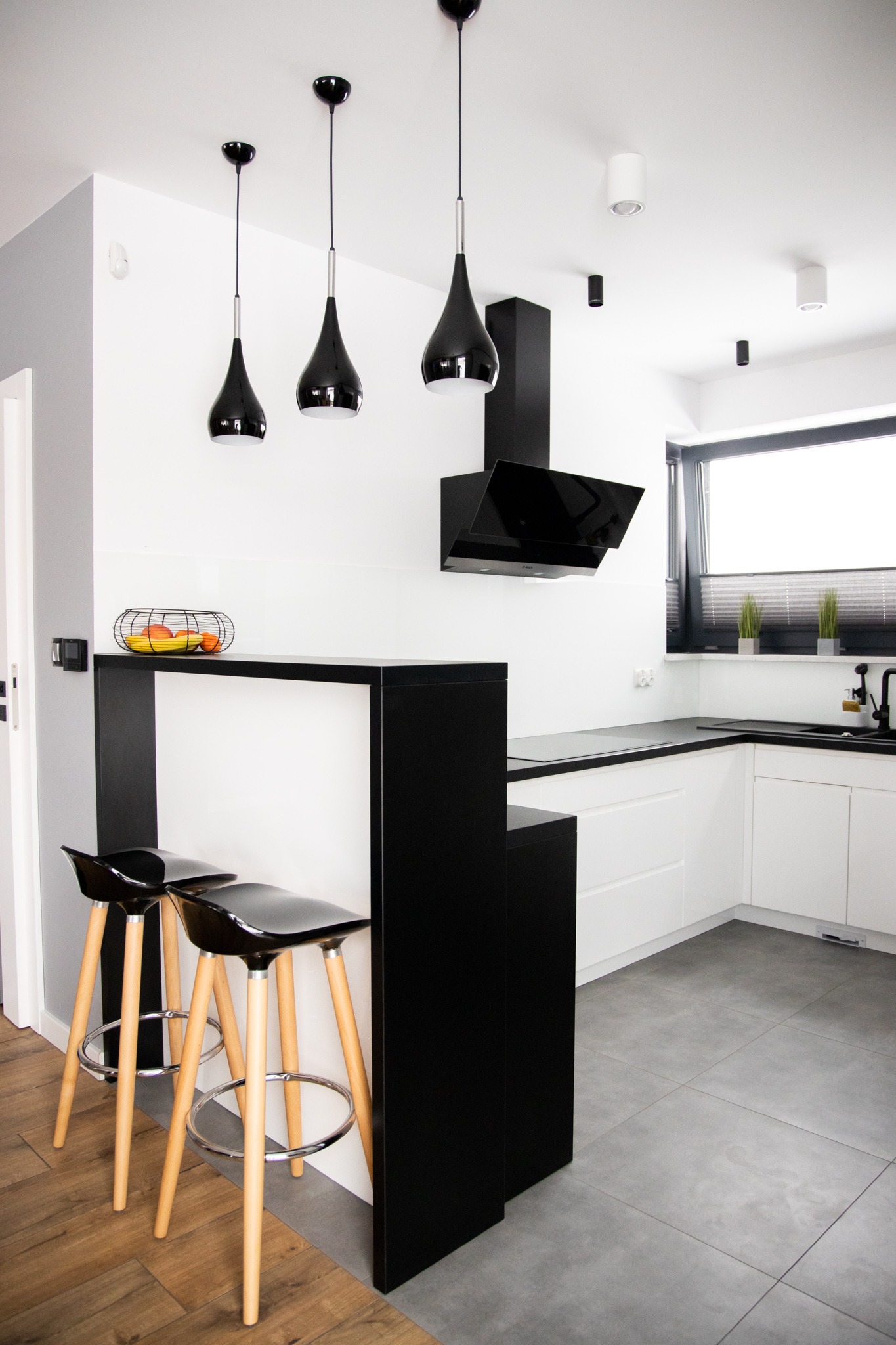 Nowoczesna kuchnia w bieli i czerni z drewnianymi akcentami: wyspa z czarnym blatem i hokerami, białe szafki, czarny okap i wiszące lampy.