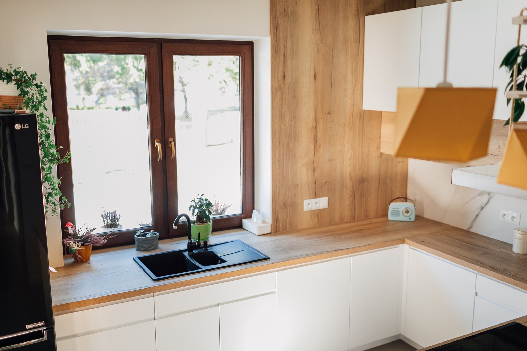 Jasna kuchnia z drewnianym blatem, czarnym zlewem pod oknem i białymi szafkami, uzupełniona o rośliny w doniczkach i retro radio.