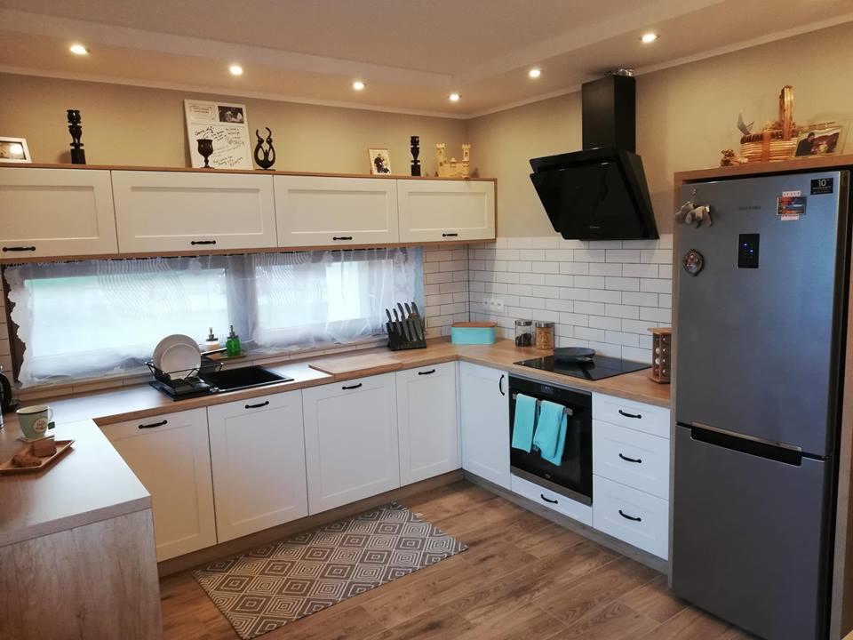 Jasna kuchnia z białymi szafkami i drewnianymi blatami, czarnym okapem i srebrną lodówką. Nad szafkami wiszącymi dekoracje, a na blacie pod oknem suszarka do naczyń.