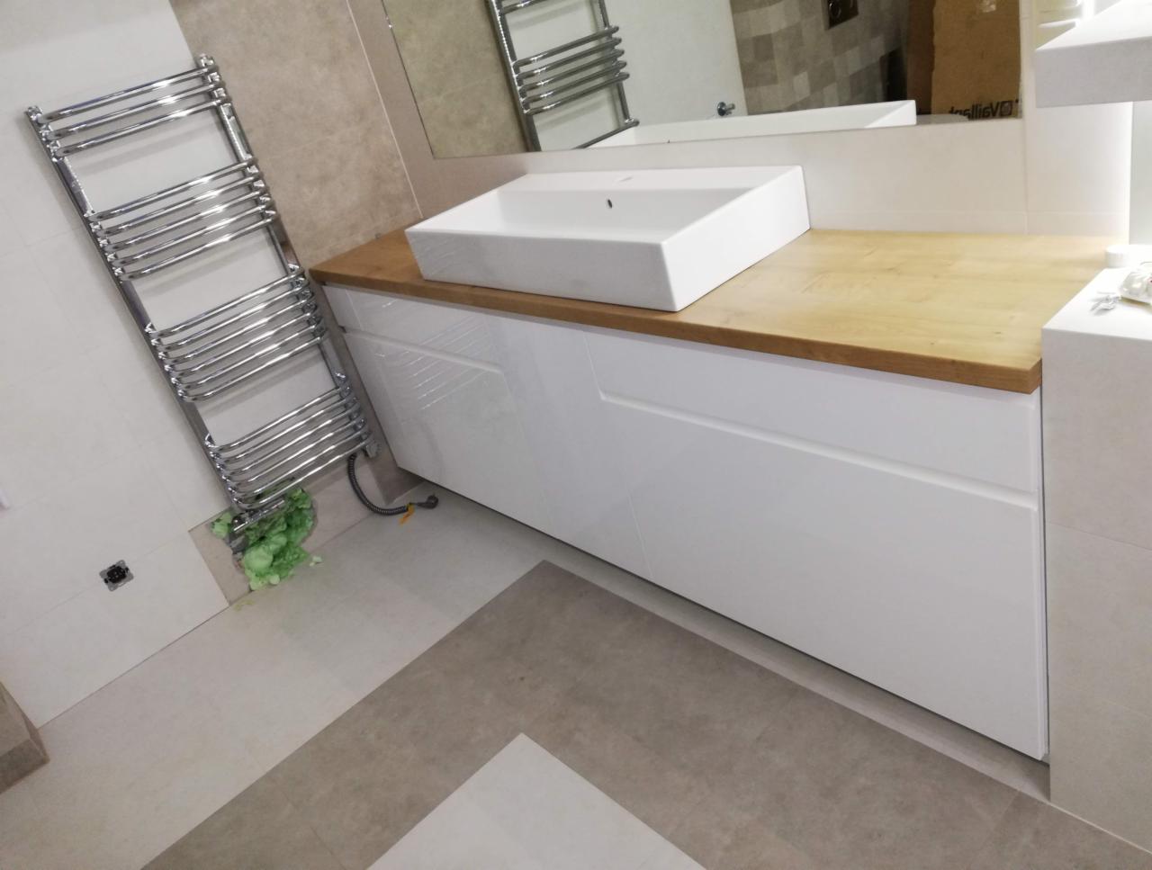 Biała szafka łazienkowa z drewnianym blatem, prostokątna umywalka nablatowa, chromowany grzejnik drabinkowy, szara podłoga z geometrycznym wzorem, widoczne ślady pianki montażowej na podłodze.