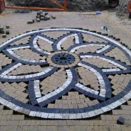 Ozdobny wzór kwiatowy z kostki brukowej w trakcie układania, widoczne niedokończone krawędzie i zapas kostki, ceglana brama w tle.