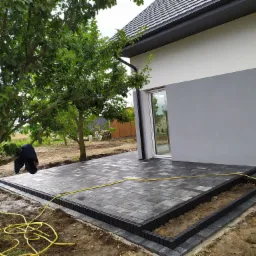 Nowo ułożony taras z kostki brukowej w odcieniach szarości, przylegający do nowoczesnego domu z szarą elewacją, z żółtym wężem ogrodowym leżącym na ziemi i bluzą wiszącą na drzewie w tle.