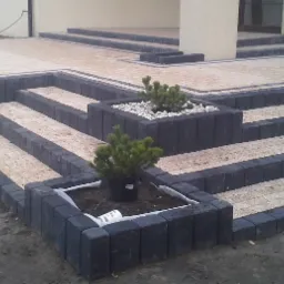 Schody zewnętrzne i taras z kostki brukowej w odcieniach beżu i grafitu, z wyeksponowanymi donicami z roślinami iglastymi i białym żwirem, prowadzące do jasnego budynku.