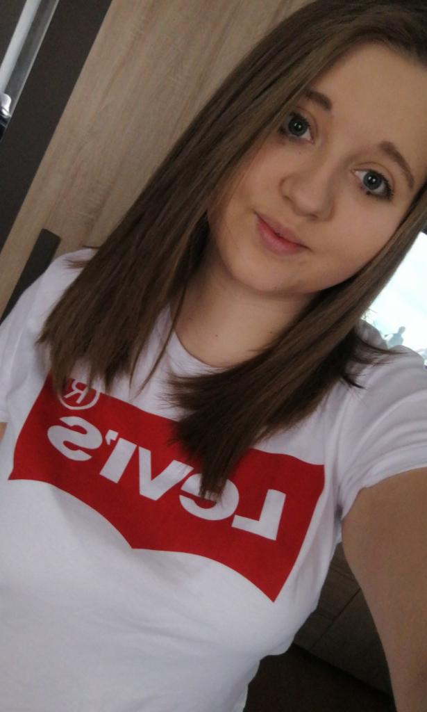 Portret młodej kobiety w białej koszulce z odwróconym czerwonym logo Levi's na tle drewnianej ściany.