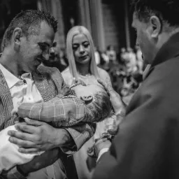 Czarno-białe zdjęcie z ceremonii chrztu: ojciec w kraciastej marynarce trzyma dziecko, ksiądz polewa wodą, w tle matka dziecka.