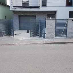 Nowoczesna, szara brama posesyjna z poziomymi panelami, częściowo otwarta, na tle domu z garażem, widoczne narzędzia i przewody sugerujące montaż w toku.