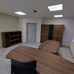 Nowoczesne biuro z dużym, drewnianym stołem konferencyjnym o nieregularnym kształcie, czarnym krzesłem biurowym, regałem z ciemnego drewna i szafą w tle, oświetlone kwadratowymi lampami sufitowymi.