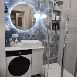 KLYMONTE - Nowoczesna łazienka z kabiną prysznicową, umywalką nablatową i okrągłym lustrem z podświetleniem LED. Na ścianach szare płytki imitujące marmur.