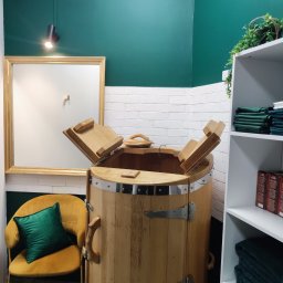 KLYMONTE - Drewniana beczka z otwartym wiekiem w pomieszczeniu z zielonymi ścianami i białymi cegłami. Obok stoi żółty fotel z zieloną poduszką i biały regał z ręcznikami.