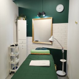 KLYMONTE - Wnętrze gabinetu kosmetycznego: zielony wystrój, łóżko do zabiegów z ręcznikiem, lustro w złotej ramie, lampa, szafka i zegar na ścianie. Stonowana kolorystyka.