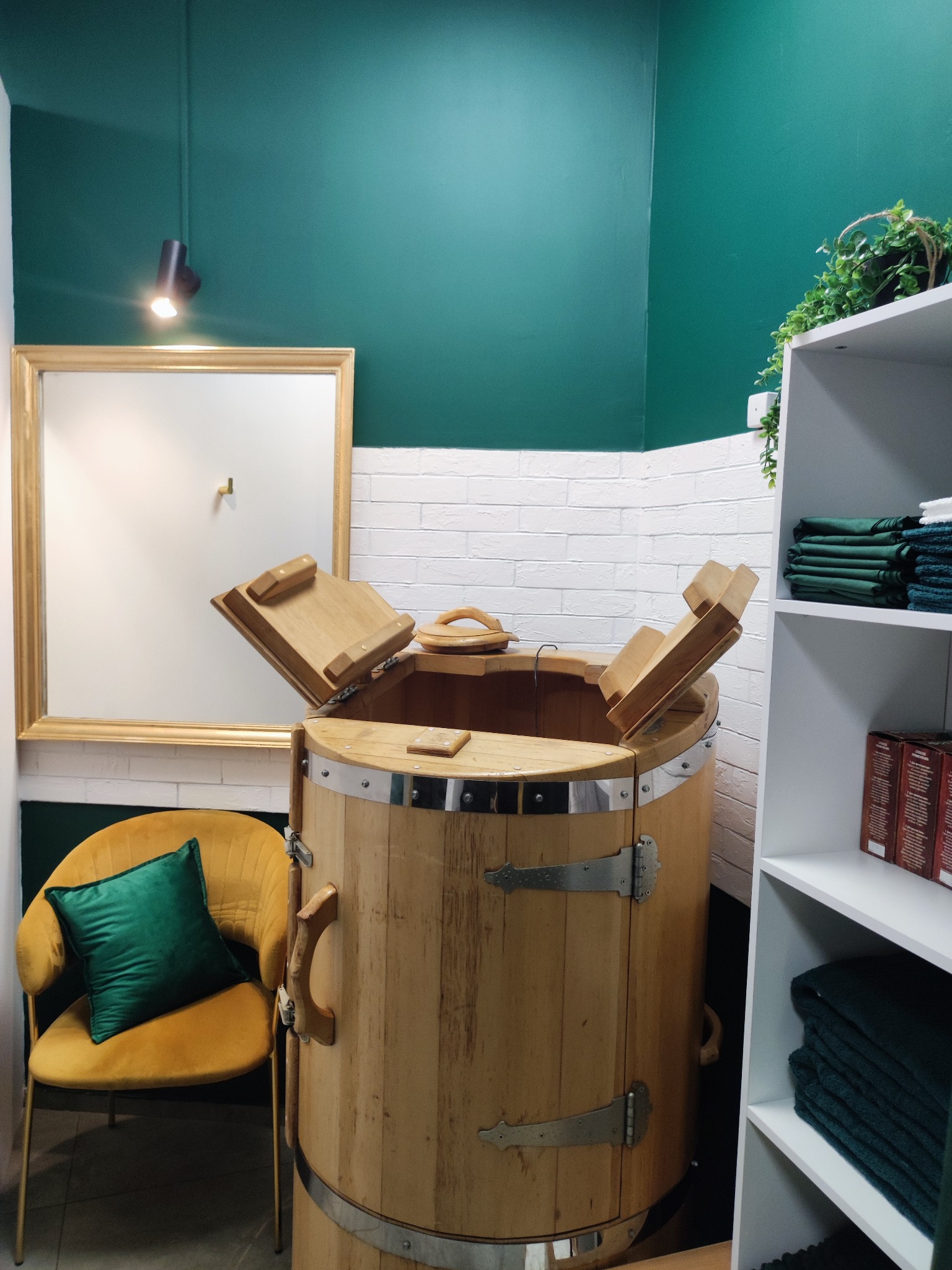 Drewniana beczka z otwartym wiekiem w pomieszczeniu z zielonymi ścianami i białymi cegłami. Obok stoi żółty fotel z zieloną poduszką i biały regał z ręcznikami.