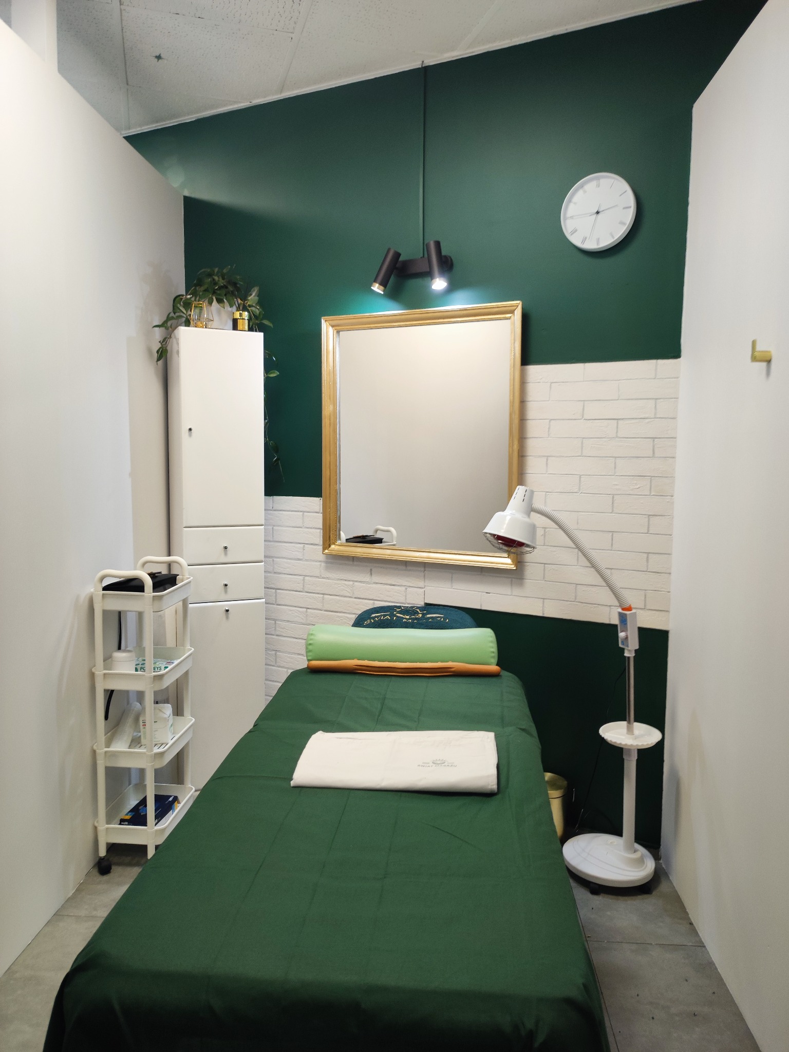 Wnętrze gabinetu kosmetycznego: zielony wystrój, łóżko do zabiegów z ręcznikiem, lustro w złotej ramie, lampa, szafka i zegar na ścianie. Stonowana kolorystyka.