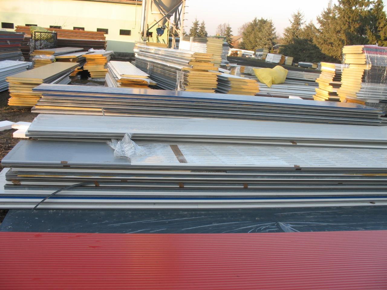 Stosy różnorodnych paneli budowlanych na zewnątrz, widoczne panele warstwowe z rdzeniem izolacyjnym, blachy trapezowe w kolorze czerwonym na pierwszym planie, w tle drzewa i silos.