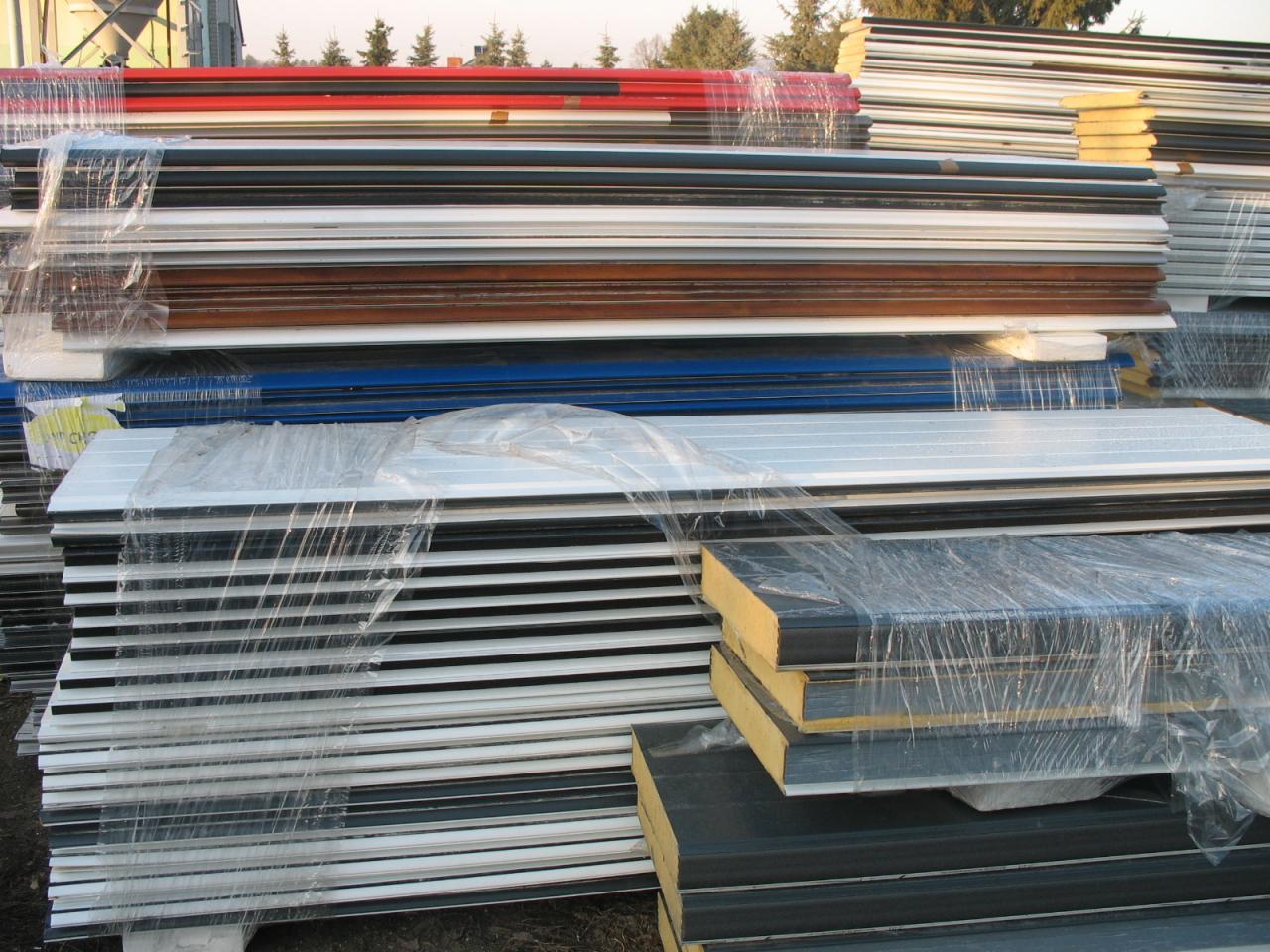 Stosy płyt warstwowych różnych kolorów (czerwony, biały, brązowy, niebieski, czarny) zabezpieczonych folią, składowane na placu budowy.