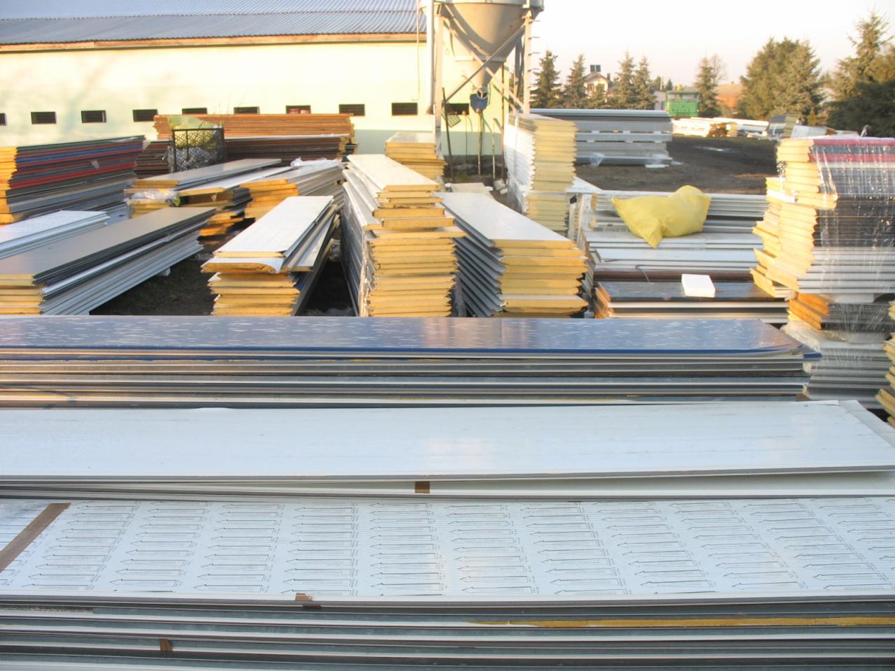 Stosy różnorodnych paneli izolacyjnych i blach na placu budowy, widoczne różne grubości i kolory, w tle budynek magazynowy i silos.