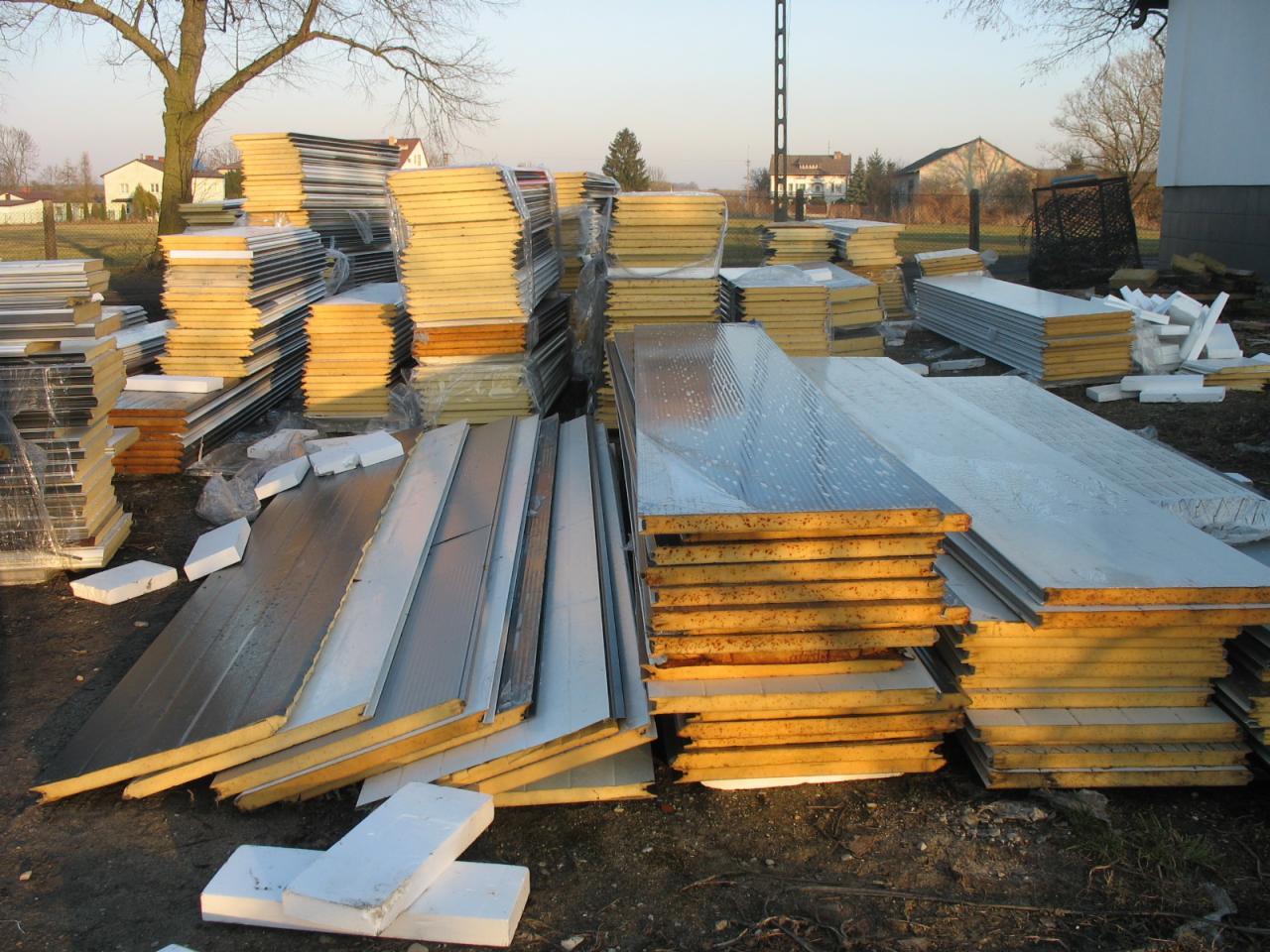 Stosy płyt warstwowych z rdzeniem poliuretanowym i metalowym wykończeniem, składowane na placu budowy w promieniach zachodzącego słońca.