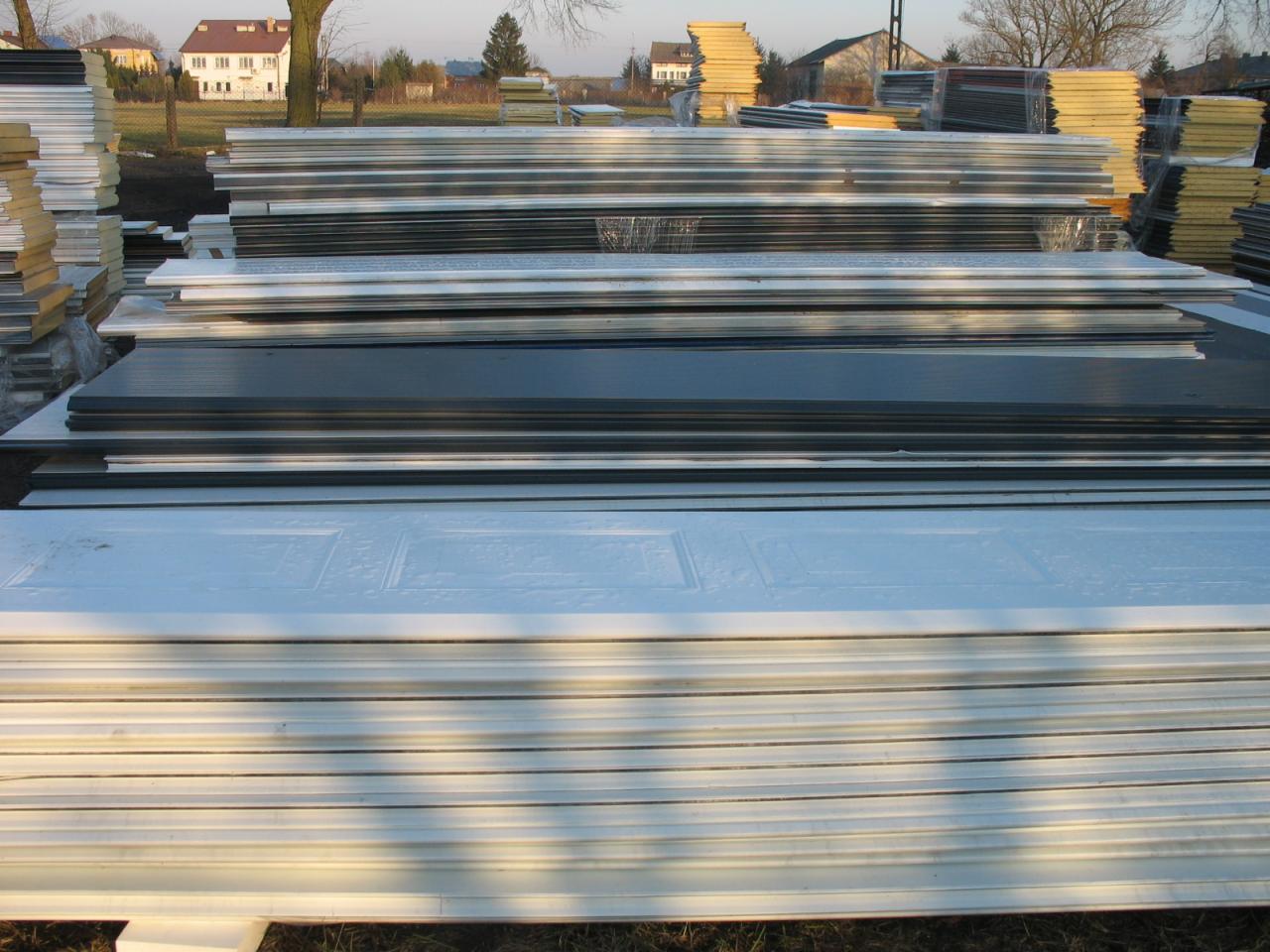 Stosy różnych płyt budowlanych, w tym białe płyty z wytłoczonym wzorem, składowane na zewnątrz na tle domów i drzew.