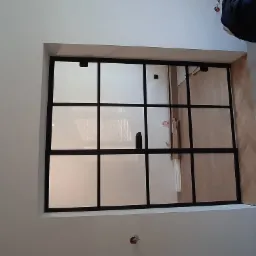 Wnętrze z zamontowaną, przeszkloną ścianką działową z czarnymi szprosami, widoczny fragment podłogi z parkietu ułożonego w jodełkę oraz niedokończone ściany.