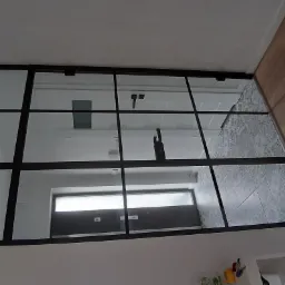 Szklana ścianka działowa z czarnymi profilami oddzielająca przedpokój z drewnianą podłogą od łazienki wykończonej płytkami w geometryczne wzory.