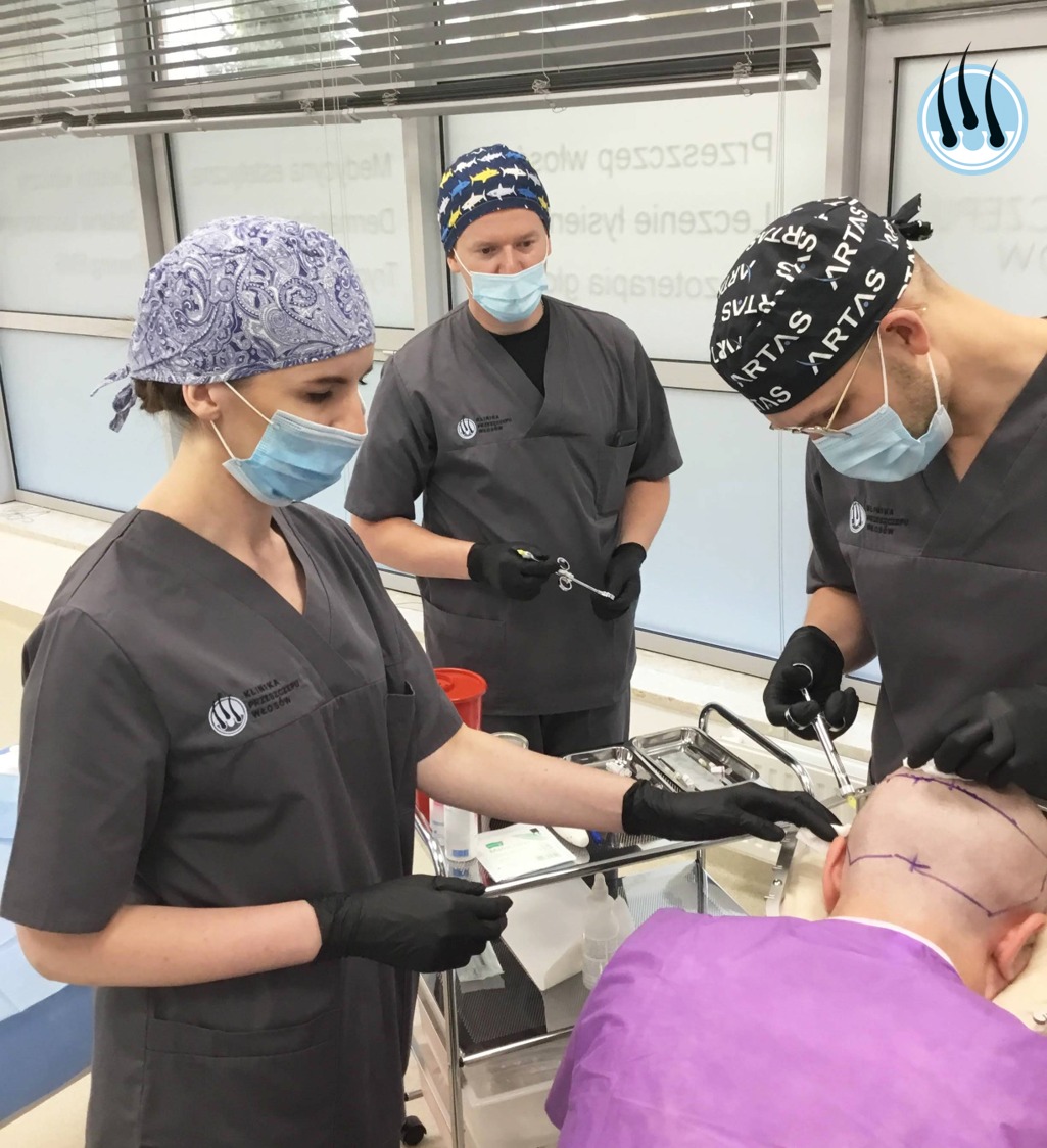 Zespół medyczny w szarych uniformach i czarnych rękawiczkach przeprowadza zabieg przeszczepu włosów pacjentowi leżącemu na stole operacyjnym, widoczne narzędzia chirurgiczne i oznaczenia na skórze...