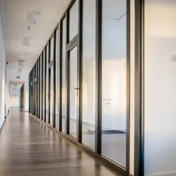 Długi korytarz biurowy z ciemnobrązową podłogą, białymi ścianami i rzędem przeszkleń w czarnych ramach, oświetlony światłem dziennym wpadającym z lewej strony.