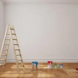 Pomalowane na biało wnętrze z drewnianą drabiną, puszkami farb i wałkiem do malowania na podłodze. Widoczne okno z białymi ramami i grzejnik.