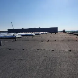 Rozległy, płaski dach pokryty ciemną papą z widocznymi łączeniami, licznymi świetlikami i elementami wentylacyjnymi; w tle budynek przemysłowy i błękitne niebo.