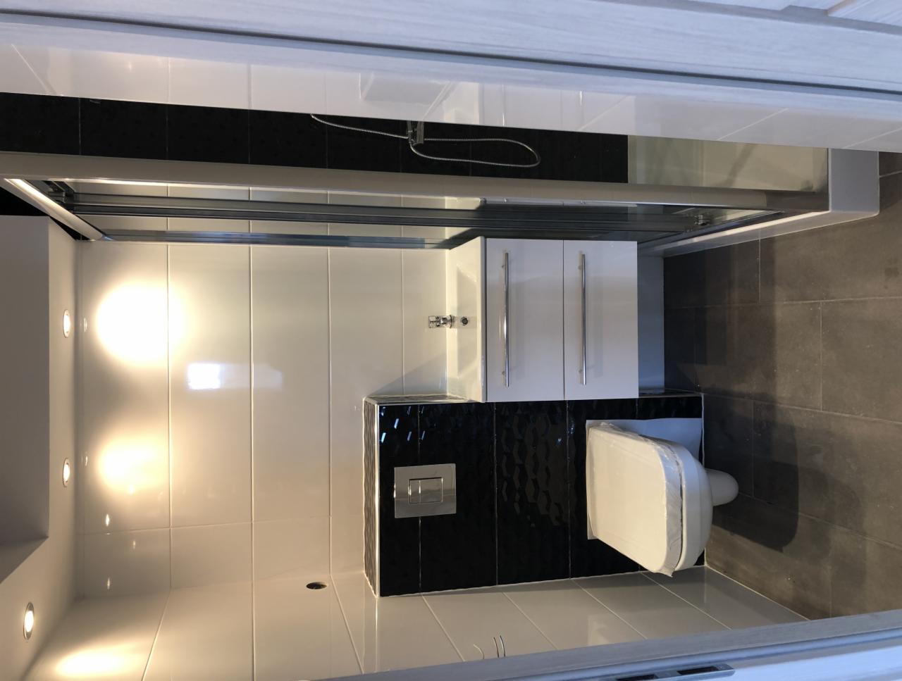 Nowoczesna łazienka z białymi płytkami ściennymi, czarnym akcentem z mozaiki za toaletą, szarą podłogą, kabiną prysznicową i białą szafką pod umywalką. Toaleta w folii ochronnej.