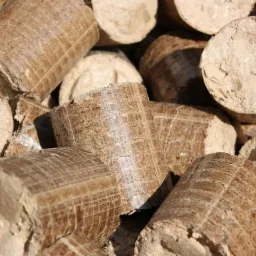 Stos brązowych brykietów z trocin o cylindrycznym kształcie, ułożonych blisko siebie pod ostrym kątem, z widoczną fakturą drewna i drobnymi pęknięciami na powierzchni.