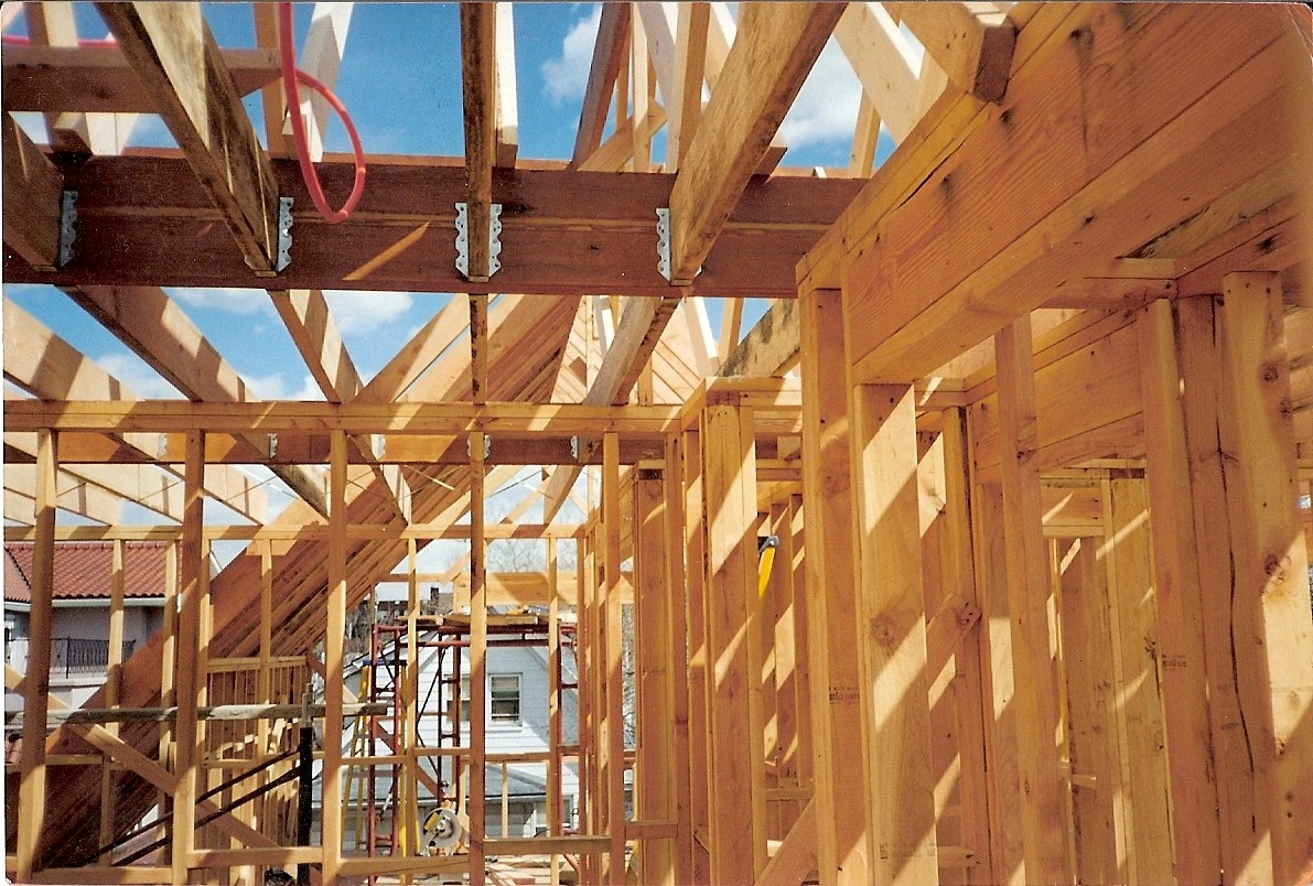 Wnętrze domu szkieletowego w trakcie budowy, widoczne drewniane elementy konstrukcyjne, złącza metalowe, czerwony kabel i fragment niebieskiego nieba przez otwory w dachu.