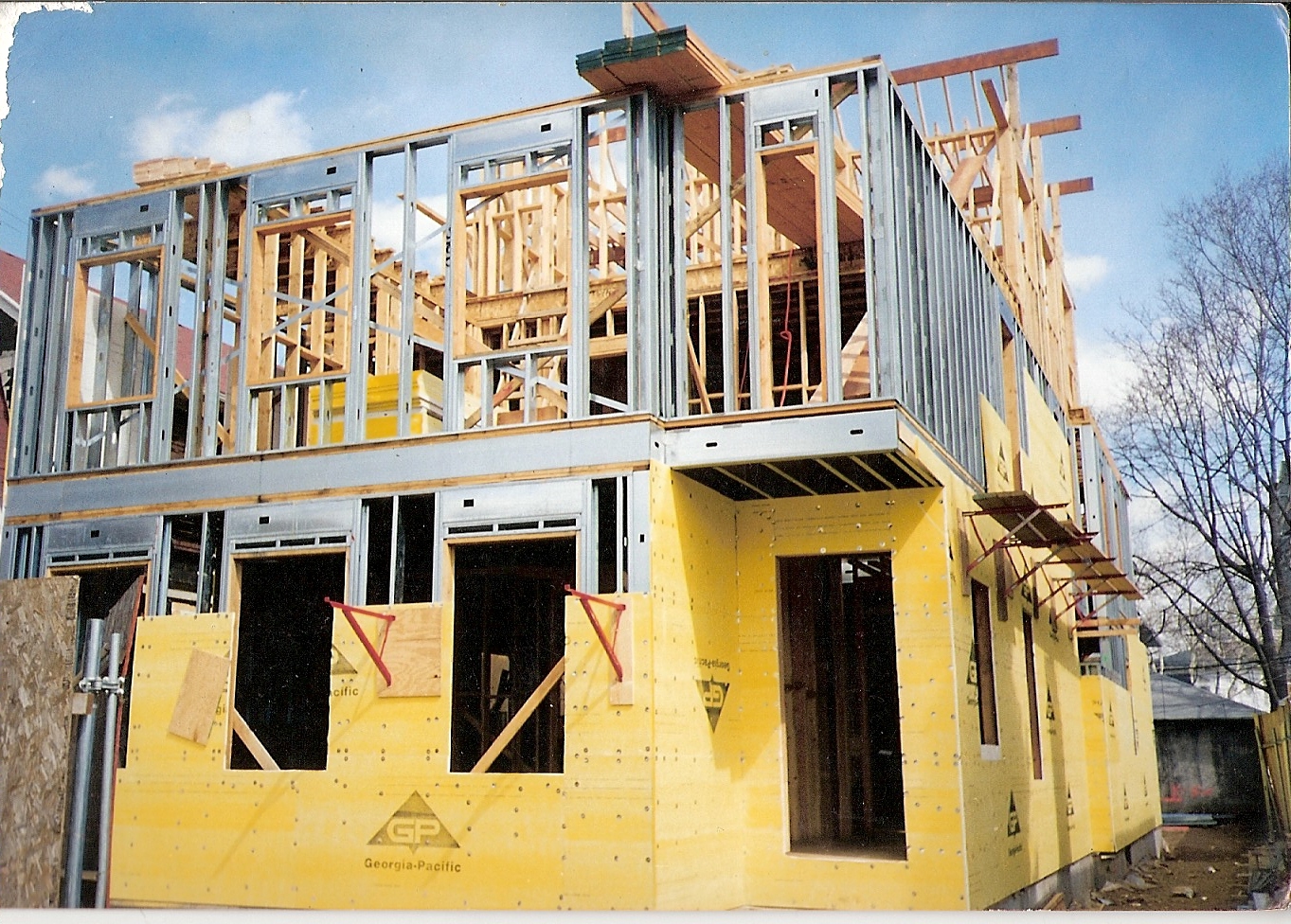 Dwukondygnacyjny dom w trakcie budowy, widoczna konstrukcja szkieletowa z drewna i metalu, żółte płyty elewacyjne na parterze, niebieskie niebo w tle.