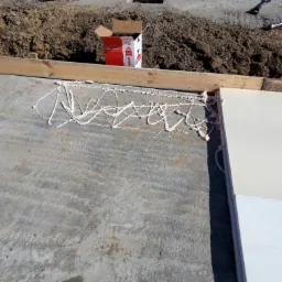 Przygotowanie podłoża pod budowę: betonowa płyta z naniesioną pianą montażową, widoczna skrzynka z pianą, narzędzia budowlane i ziemia w tle.