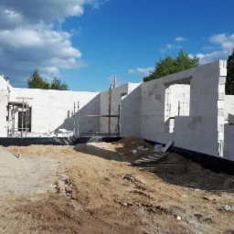 Wznoszenie ścian zewnętrznych domu z białych bloczków, widoczne rusztowania, przygotowany teren budowy i fragment sąsiedniego domu.