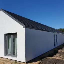 Nowoczesny, parterowy dom z białymi ścianami i ciemną, spadzistą dachówką, widoczny z zewnątrz w słoneczny dzień, z przygotowanym terenem wokół.